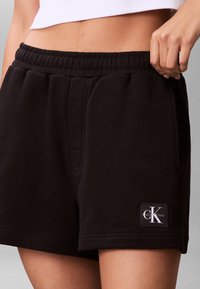 Person iført sorte Calvin Klein shorts med elastisk talje og en hvid cropped top, hånd hvilende nær lomme på højre side.