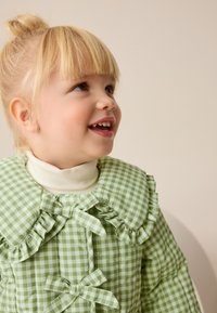 Grünes kariertes Kleid mit gerafftem Kragen und Schleifendetails, mit einer gesteppte Textur. Gestylt mit einem weißen Rollkragenpullover darunter.