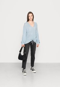 Suéter de malha canelada azul claro com decote em V e bainha com cordão. Combinado com jeans escuros e ténis pretos; segurando uma mala de ombro preta.