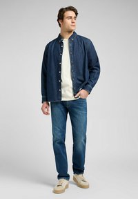 Camisa de denim en azul oscuro, estilo abotonado con un collar clásico sobre una camiseta blanca. Combinada con jeans azul oscuro y zapatillas beige.
