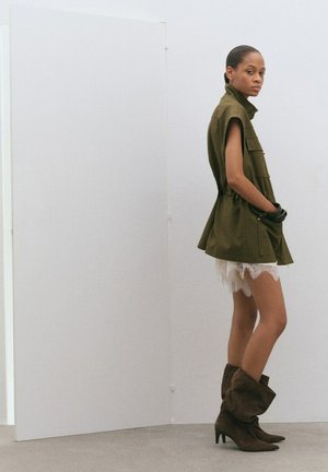 Femme debout de profil portant une veste sans manches vert olive, un short en dentelle blanche et des bottes marron à talons tombantes, sur un fond uni.