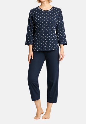 Frau trägt ein dunkelblaues Pyjama-Set mit kleinen weißen Blumenmustern auf dem Oberteil und einfarbige Hose, steht barfuß vor weißem Hintergrund.