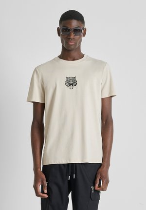 Muškarac u bež majici s crnim grafikonom tigra i crnim hlačama s vezicama, stoji nasuprot jednostavnoj svijetloj pozadini.