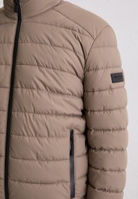 Beige gewatteerd jack met horizontale stiksels, hoge kraag en een zak met ritssluiting. Voorzien van een zwarte rits en een label van textiel met structuur.