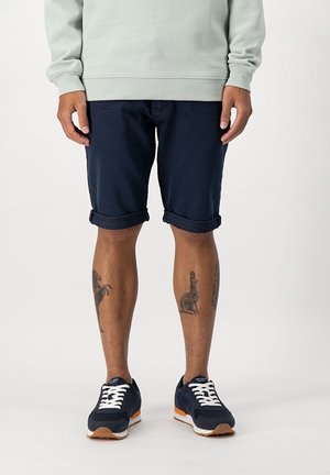 Person iført mørkeblå shorts med opklemmet kant, mørkeblå sneakers med hvide snørebånd og en lysegrøn sweatshirt, stående foran en ensfarvet baggrund.