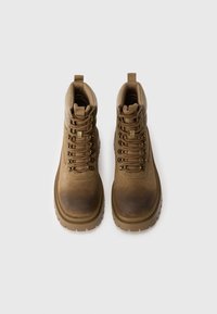 Botas altas marrones con un acabado de ante texturizado, sistema de cordones, cuello acolchado y suela de goma gruesa. Presenta acentos de herrajes metálicos.
