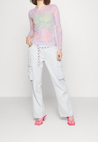 Transparente Langarmbluse mit einem mehrfarbigen Wellenmuster, kombiniert mit hellgrauen Cargo-Hosen mit Utility-Taschen. Rosa Riemchen-Absätze.