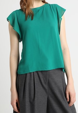 Blusa teal a maniche corte realizzata in tessuto leggero con scollatura rotonda e spalle leggermente drappeggiate, abbinata a pantaloni grigi a gamba larga.
