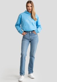 Sudadera azul cielo con un bolsillo frontal y texto de logo blanco, combinada con jeans de tiro alto en un azul claro y zapatillas blancas. Tela suave, estilo casual.