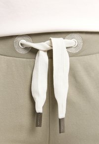 Khaki-Jogginghosen mit strukturierter Oberfläche, ausgestattet mit einer weißen, flachen Kordel mit Metallenden und runden Ösen.