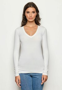 Weißes langärmliges Oberteil mit einem tiefen V-Ausschnitt, das eine figurbetonte Passform und einen weichen Stoff aufweist, kombiniert mit blauer Jeans und somit einen lässigen Look schafft.