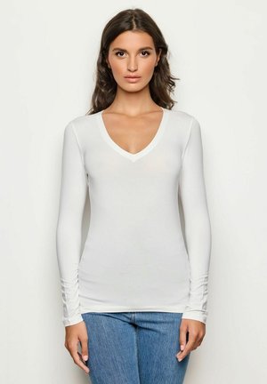 Top bianco a maniche lunghe con scollatura a V profonda, caratterizzato da un design aderente e una texture morbida, abbinato a jeans blu, creando un look casual.