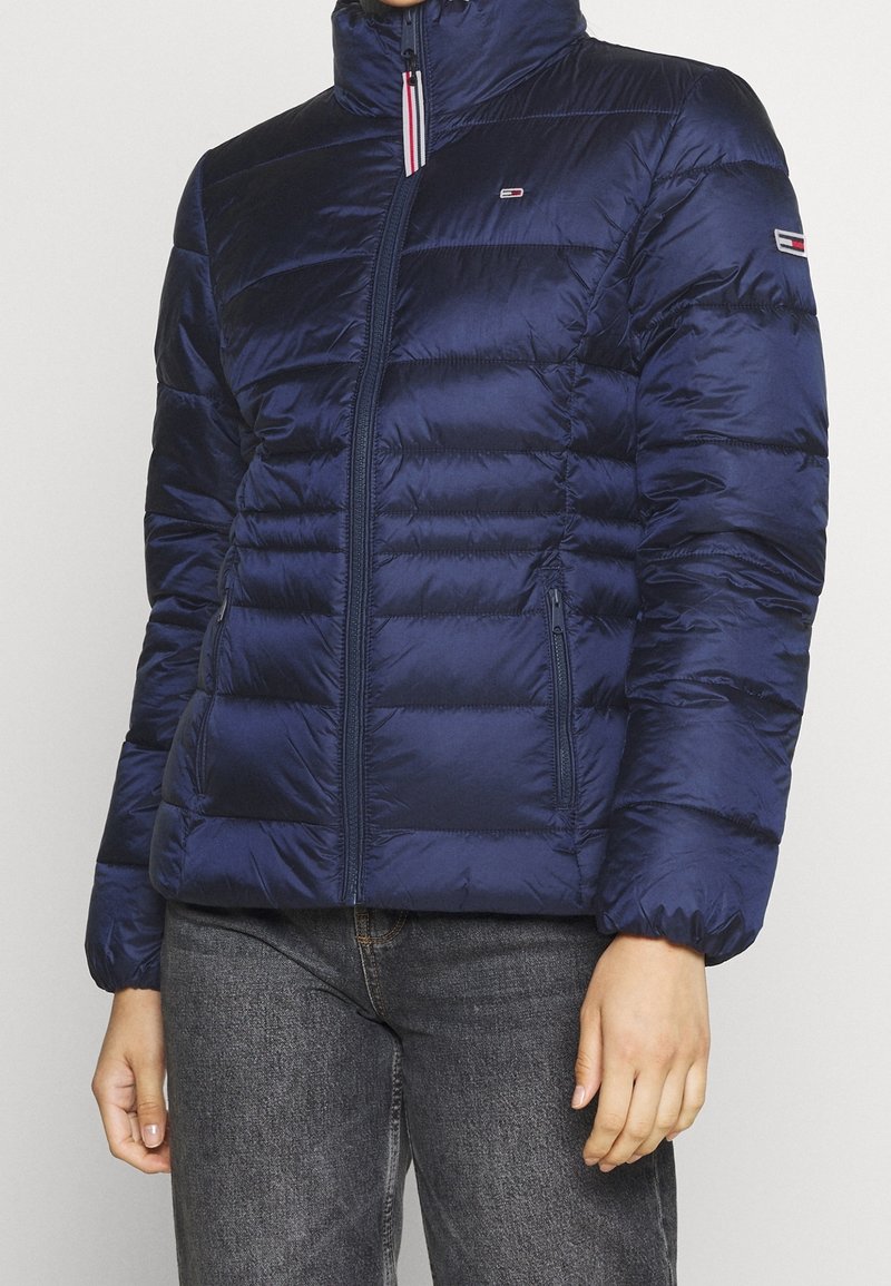 Giacca a vento imbottita blu navy con collo alto, chiusura a zip, tasche laterali e cuciture orizzontali sottili. Presenta il logo sul petto e sulla manica.