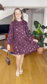 Rochie mov cu embellisheuri florale și accente din mărgeluțe, mâneci lungi, lungime deasupra genunchilor, asortată cu sandale albe și o geantă mică.