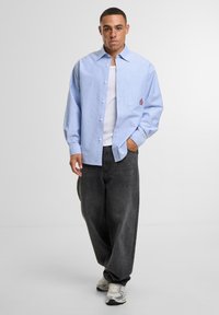 Light blue striped button-up shirt with long sleeves, paired with dark baggy jeans and white sneakers. Features a pocket with a red logo.