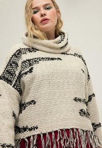Chunky gestrickter Pullover in Creme mit schwarzen abstrakten Mustern, einem Cowl-Neck und Fransen entlang dem Saum. Weicher Stoff, lockere Passform.