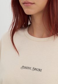 Camiseta de algodón beige con cuello redondo y texto negro "POSITIVE SINCERE". Textura suave con un sutil detalle de costura.