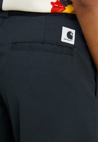 Fekete szövetnadrág hátsó zsebbel, fehér Carhartt logó címkével. A felület simának és tartósnak tűnik.