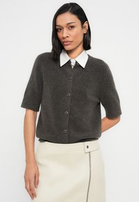 Καφέ καρό μπλουζάκι με κοντά μανίκια και κουμπιά μπροστά, κατασκευασμένο από μαλακό υφασμάτινο ύφασμα, συνδυασμένο με λευκό πουκάμισο με γιακά και κρεμ δερμάτινη φούστα.