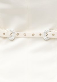 Λευκή υφασμάτινη ζώνη με διπλές διακοσμητικές αγκράφες και ασημένια ελάσματα σε κρεμ χρωματιστό υφασμάτινο ένδυμα με υφή.