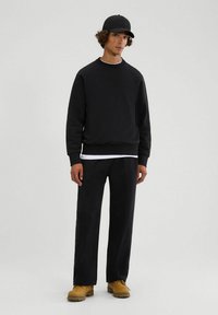 Schwarzer Hoodie mit gerippten Bündchen, über einem weißen Hemd getragen; schwarze Hose und gelbe Arbeitsschuhe. Das Modell trägt eine passende schwarze Mütze.