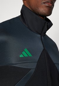 Veste zippée noire et gris foncé avec un col montant, texture lisse et un logo vert à trois rayures bien visible sur la poitrine.