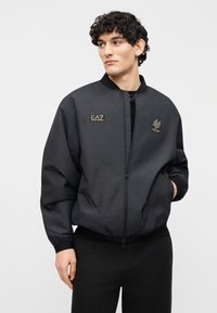 Άνδρας που φοράει μαύρο μπουφάν bomber με φερμουάρ, με λογότυπα EA7 και Milano Italia, στέκεται μπροστά σε απλό φωτεινό φόντο, με το αριστερό χέρι στην τσέπη.