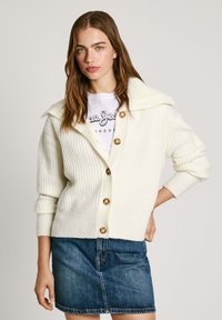 Creme gekleurde gebreide cardigan met ribbelstructuur, grote kraag en bruine knopen. Gecombineerd met een casual wit t-shirt en een spijkerrok.
