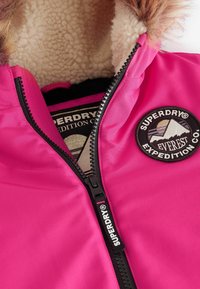 Veste rose avec capuche à fourrure, fermeture éclair, et un patch rond représentant des montagnes. Intérieur doublé de douce polaire blanche.