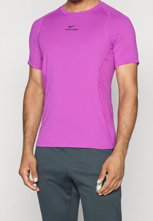 Mężczyzna w dopasowanej, jaskrawo fioletowej sportowej koszulce Nike Pro z krótkimi rękawami i ciemnoszarych spodniach, stojący ze zrelaksowanymi rękami.