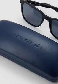 Ochelari de soare negri cu lentile întunecate alături de o carcasă rigidă maro închis, având un logo "LACOSTE" embosat și un emblema cu un crocodil.