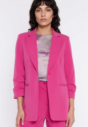 Kobieta w jaskraworóżowej marynarce z podwiniętymi rękawami, na wzorzystej purpurowo-szarej bluzce, w połączeniu z pasującymi różowymi spodniami.