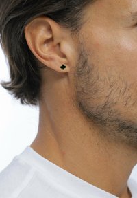 Seitenansicht eines Mannes mit kurzen Haaren, der einen kleinen schwarzen und goldenen Ohrstecker in Form eines Kleeblatts und spärlichen Gesichtshaaren trägt, bekleidet mit einem weißen Hemd.