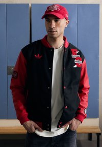 Giacca varsity nera e rossa con toppe, inclusa "BAYERN MONACO," e un logo ricamato Adidas. Indossata sopra una maglietta bianca e jeans.