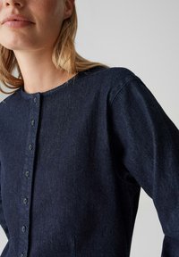 Dunkelblaues Denim-Kleidungsstück mit rundem Ausschnitt, Knopfleiste vorne, langen Ärmeln und Seitentaschen. Der Stoff wirkt strukturiert mit einem dezenten Glanz.
