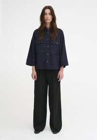 Giacca di jeans di un blu scuro con due tasche sul petto, chiusura a bottoni e maniche ampie, abbinata a pantaloni neri a gamba larga e scarpe basse.