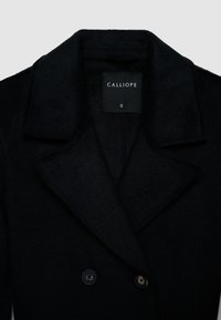 Cappotto nero doppiopetto con colletto sagomato e due bottoni. Il tessuto presenta una superficie morbida e testurizzata. Etichetta con la scritta "CALLIOPE," taglia S.