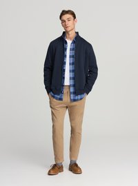 Giacca con zip blu navy sopra una camicia a quadri blu, maglietta bianca, pantaloni kaki e scarpe marroni con lacci strutturati.