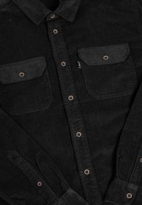 Schwarzes Cordhemd mit kupferfarbenen Knöpfen, zwei Brusttaschen, spitzem Kragen und Knopfmanschetten, aus einem weichen, strukturierten Stoff gefertigt.