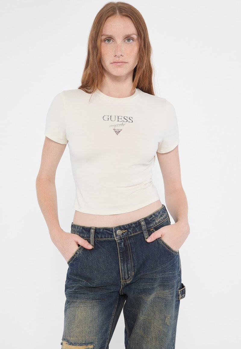 Top corto de manga corta en color crema con el logo "GUESS" en letras metálicas, combinado con unos jeans de mezclilla oscuros de tiro alto y desgastados.