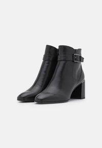 Botas de tobillo de cuero negro con punta redonda, tacón cuadrado de altura media y detalle de hebilla en la correa del puño. Textura suave.