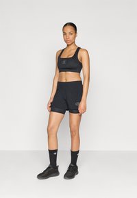 Conjunto de sujetador deportivo y pantalones cortos negros, con un logo. El material es suave y tiene un diseño ajustado. Zapatillas negras y calcetines completan el look.