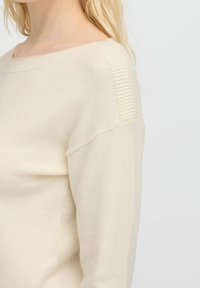 Maglione beige lavorato a maglia con un ampio scollo a barca, dettagli a costine sulle cuciture delle spalle e un tessuto morbido e strutturato. Design semplice e versatile.