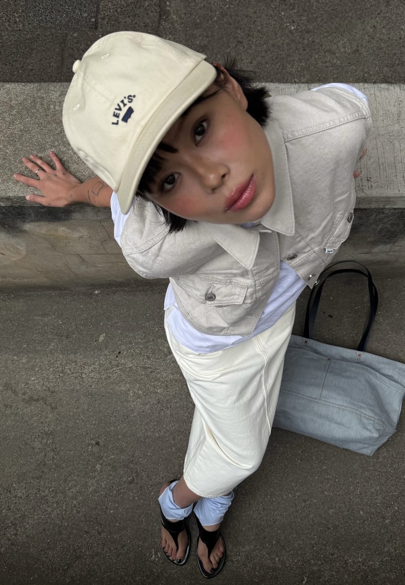 Căciulă bej cu logo-ul Levi's, vestă ușoară bej, tricou alb, fustă crem, sandale negre, geantă tote cu dungi pe partea laterală, fundal gri.