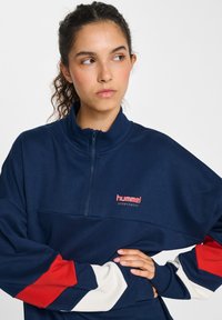 Felpa con zip di colore blu navy con accenti a blocchi di colore rosso, bianco e crema sulle maniche. Presenta un piccolo logo sul petto e un collo alto.