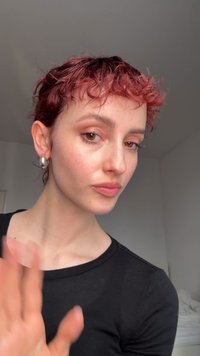 Personne aux cheveux roux courts et bouclés, portant des boucles d'oreilles anneaux en argent et un haut noir, agitant la main dans une pièce lumineuse aux murs blancs.