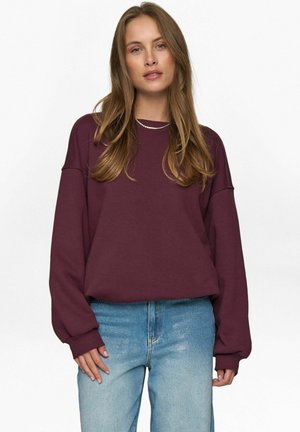 Bordeauxfarbenes Sweatshirt mit langen Ärmeln und Rundhalsausschnitt; kombiniert mit hellblauer Jeans. Der Stoff wirkt weich und leicht strukturiert.
