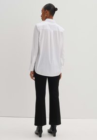 Camicia bianca con colletto classico e maniche lunghe, abbinata a pantaloni neri a zampa d'elefante e stivaletti, su uno sfondo neutro.
