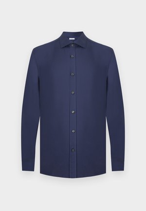 Homme aux cheveux bouclés portant une chemise bleu marine à boutons, un pantalon gris, des baskets blanches, debout avec une main dans la poche, regardant vers la droite.