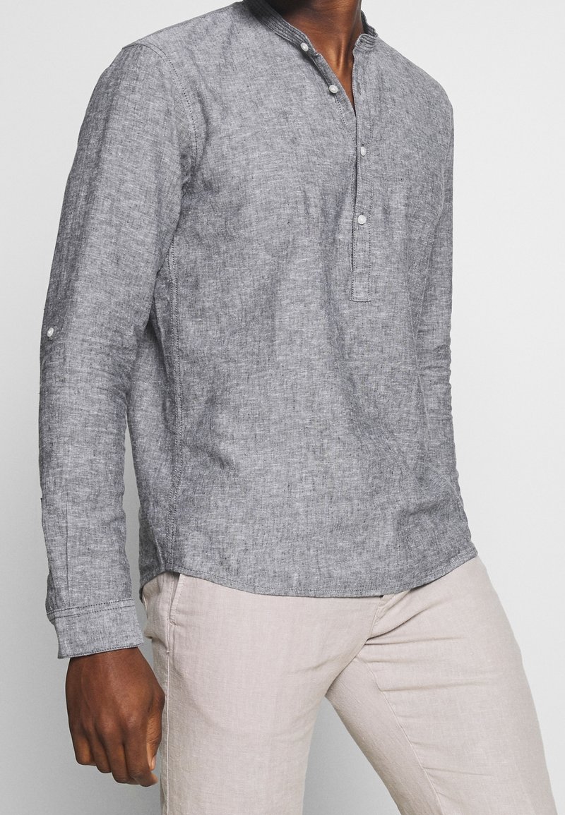 Harmaa pellavainen pitkähihainen paita, jossa on puolipainikkeellinen nappilista ja mandariinikaulus; siinä on hienovarainen tekstuuri ja käärityt hihat.